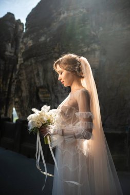 Dağların arka planında bir buket çiçekle beyaz elbiseli bir gelin ve İsviçre Saksonya, Almanya, Bastei 'de bir dağ geçidi..