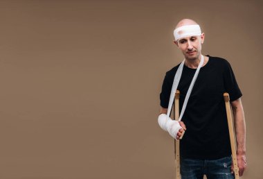 Kafası bandajlı ve kolunda alçı olan hırpalanmış bir adam gri bir arka planda koltuk değnekleriyle duruyor..