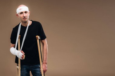 Kafası bandajlı ve kolunda alçı olan hırpalanmış bir adam gri bir arka planda koltuk değnekleriyle duruyor..