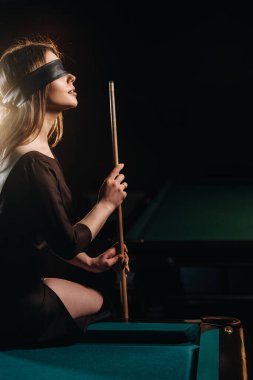 A girl with a blindfold and a cue in her hands is sitting on a table in a billiard club.Russian billiards. clipart