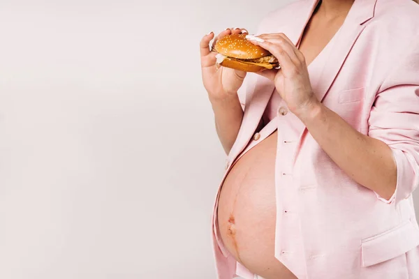 Primo piano di una ragazza incinta in giacca e cravatta con hamburger in mano su sfondo grigio — Foto Stock