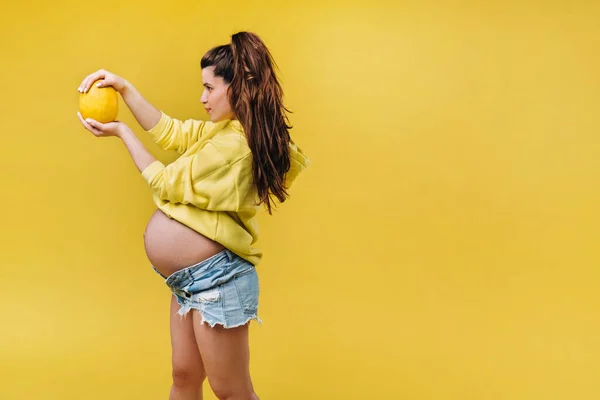 Una embarazada con una chaqueta amarilla está de pie con un melón en las manos sobre un fondo amarillo aislado — Foto de Stock
