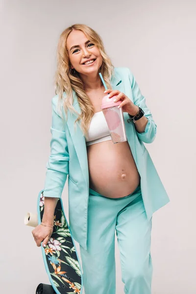 Chica embarazada en ropa turquesa con un vaso de jugo y un monopatín en sus manos sobre un fondo azul — Foto de Stock