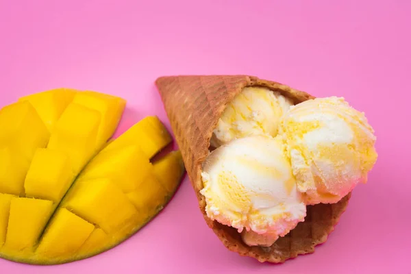 Mango-flavored ice cream balls in a waffle cone on a pink background. Ripe mango with ice cream on a pink background