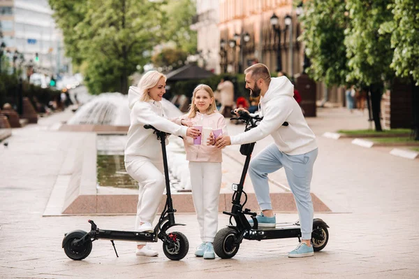 A family in white clothes stands in the city on electric scooters and drinks a drink.