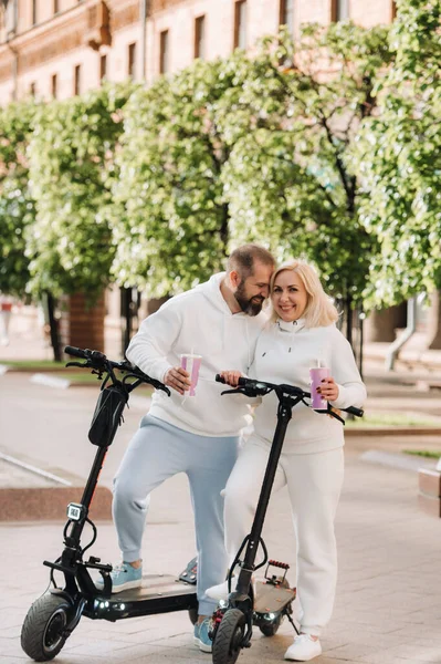 Beyaz elbiseli bir çift, şehirde elektrikli scooterlarla ellerinde içkiyle duruyorlar..