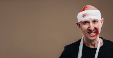 Kafası bandajlı ve kolunda alçı olan hırpalanmış bir adam gri bir arka planda duruyor..