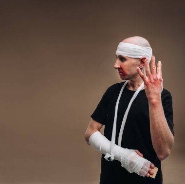 Kafası bandajlı ve kolunda alçı olan hırpalanmış bir adam gri bir arka planda duruyor..