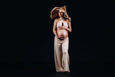 Stüdyoda siyah arka planda bej elbiseli hasır şapkalı hamile bir kadın.