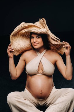 Hasır şapkalı hamile bir kadın, siyah arka plandaki stüdyoda bej giysiler içinde yerde oturuyor..