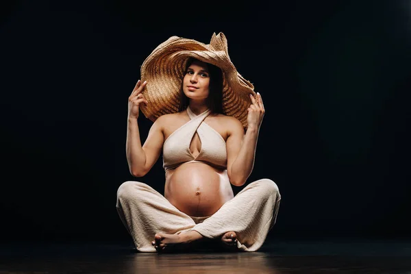 一个戴着草帽的孕妇穿着米黄色衣服坐在地板上 背景是黑色的 — 图库照片