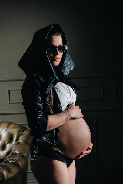 一个身穿黑色衣服 戴着眼镜 头戴头巾的孕妇站在黑漆漆的工作室里 她的肚子很大 — 图库照片