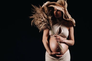 Stüdyoda elinde buket bej elbiseli, hasır şapkalı, siyah arka planda hamile bir kadın..