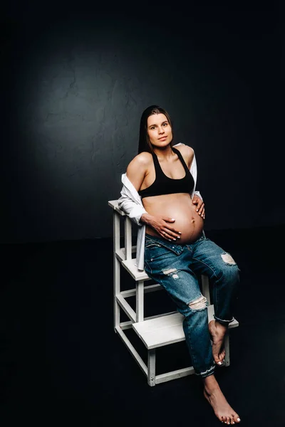 一个身披黑色背景的孕妇坐在工作室的梯子椅子上 — 图库照片
