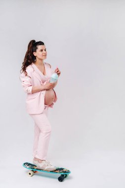 Elinde kaykayıyla pembe takım elbiseli hamile bir kız gri bir arka planda duruyor..