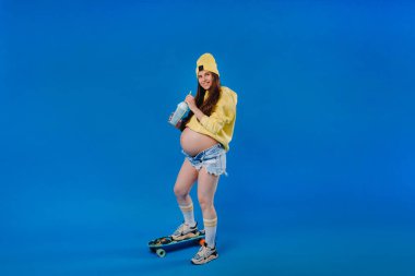 Sarı elbiseli hamile bir kız bir bardak meyve suyuyla mavi arka planda kaykaya biniyor..