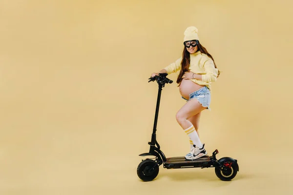 孤立した黄色の背景に電動スクーターの黄色の服の妊娠中の女の子 — ストック写真