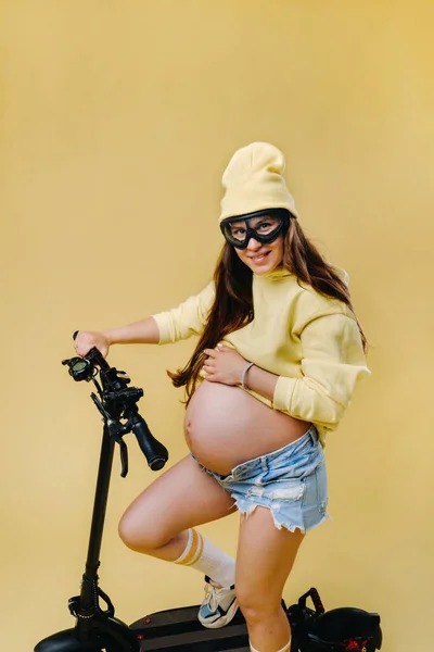 一个穿着黄色衣服的怀孕女孩骑着电动车在一个孤立的黄色背景上 — 图库照片