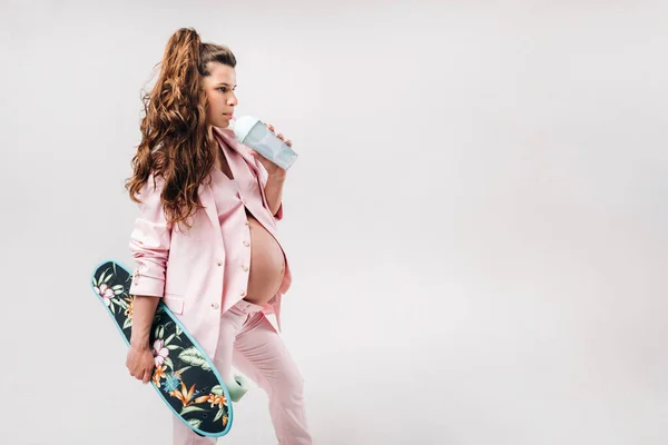 妊娠中の女の子でピンクのスーツにスケートボードで彼女の手にジュースを飲む灰色の背景 — ストック写真