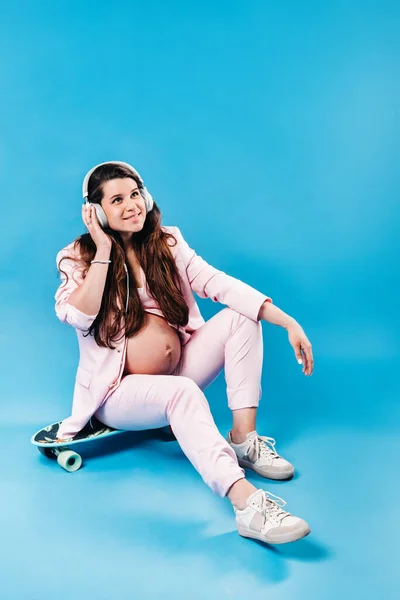ピンクのスーツを着た妊婦がヘッドフォンでスケートボードに座って青い背景で音楽を聴く — ストック写真