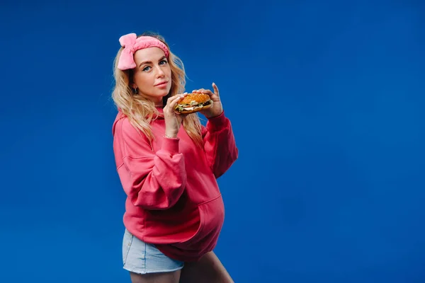 青い背景にハンバーガーを手にピンクの服を着た妊婦が — ストック写真