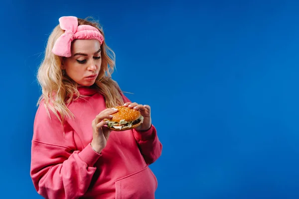 青い背景にハンバーガーを手にピンクの服を着た妊婦が — ストック写真