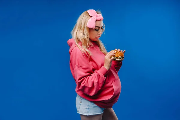 Беременная Девушка Розовой Одежде Гамбургером Руках Синем Фоне — стоковое фото