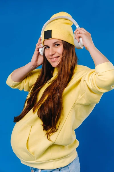 一个穿着黄色夹克和耳机的怀孕女孩站在蓝色的背景上 — 图库照片