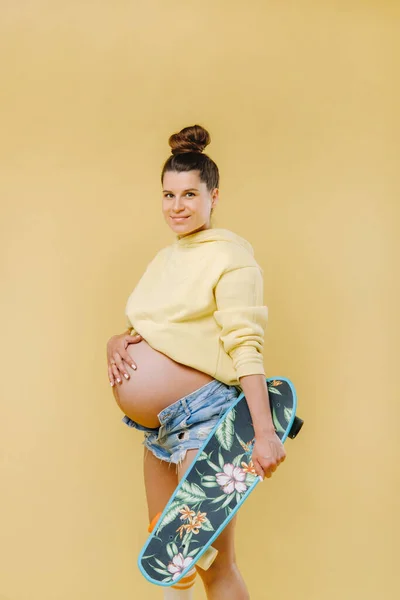 穿着黄色夹克 手里拿着滑板 背景是黄色的怀孕女孩 — 图库照片