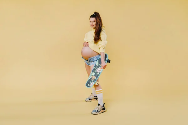 妊娠中の女の子でA黄色のジャケットとともにスケートボード上の彼女の手で黄色の背景 — ストック写真