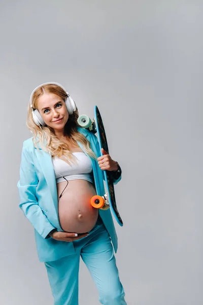 彼女の手とヘッドフォンでスケートボードとターコイズスーツの妊婦は灰色の背景に立っています — ストック写真