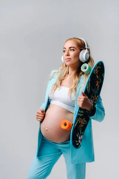 彼女の手とヘッドフォンでスケートボードとターコイズスーツの妊婦は灰色の背景に立っています — ストック写真