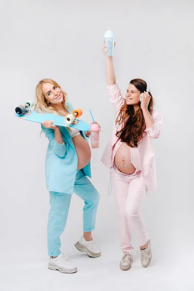 ターコイズとピンクのスーツを着た2人の妊婦ジュースのグラススケートとヘッドフォンスタンド灰色の背景 — ストック写真
