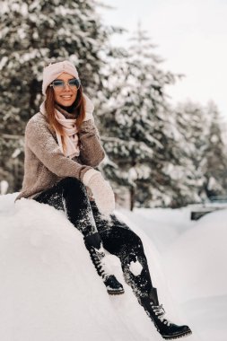 Kışın süveter ve gözlüklü bir kız ormanda karla kaplı bir arka planda oturuyor..