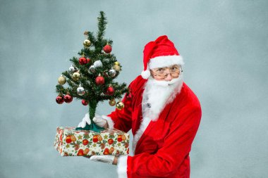 Gri arka planda Noel Baba 'nın portresi. Noel Baba 'nın elinde bir hediye ve güzel bir Noel ağacı var. Duygu kavramı.