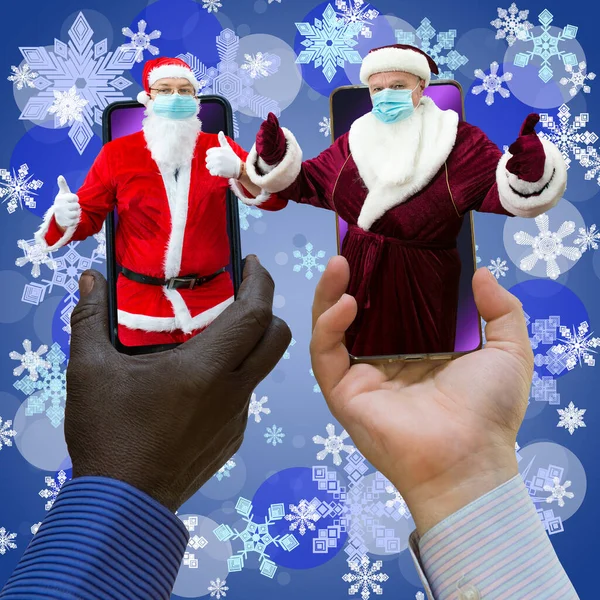 非裔美国人和白人手牵着两个圣诞老人的手机 圣诞老人戴着口罩 蓝底雪片的特写 — 图库照片