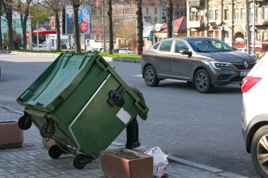 Dnepropetrovsk, Ukrayna - 04.21.2021: Sokaktaki çöp tenekesinde yiyecek arayan fakir kadın. Evsizin ne parası ne de evi var.