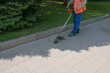 Tulumlu bir işçi elinde makasla çimleri biçer ve parktaki patikaları biçer. Toprak bakımı.
