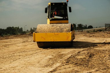 Asfalt üzerinde sarı ağır titreşimli silindir çalışıyor. Yol tamiri. Güneşli bir yaz gününde otoyol tamiri. Ağır makineler, forkliftler ve kamyonlar.