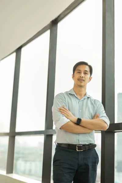 Asiático hombre de negocios de pie en su brazos cruz al lado de Grande ventana. — Foto de Stock