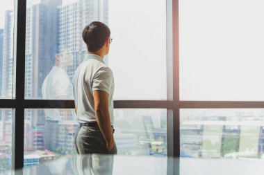 Elleri ceplerinde duran bir adam şehir binasının penceresinden dışarı bakıyor..