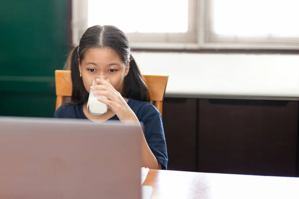 小女孩喝牛奶，在家用笔记本电脑上网学习. — 图库照片