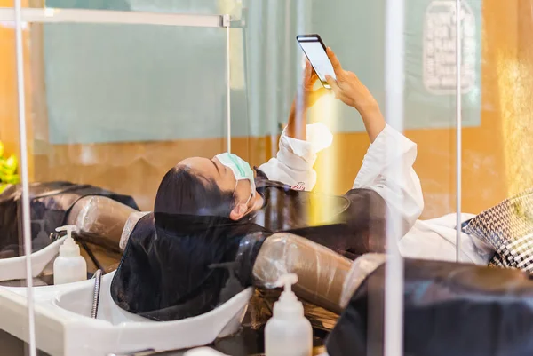 Frau in Schutzmaske mit Handy wartet auf Friseur, der Haare wäscht. — Stockfoto