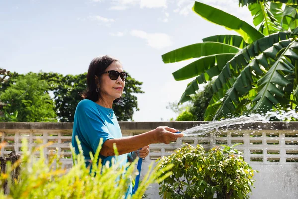 Mujer mayor regando plantas con manguera en el jardín en casa. exterior —  Fotos de Stock