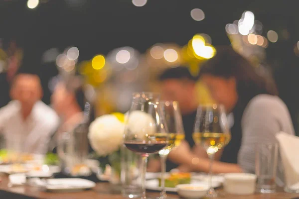 Copos para vinho e pessoas à mesa de jantar com fundo embaçado e luzes quentes. a elebrar o conceito da noite. interior — Fotografia de Stock