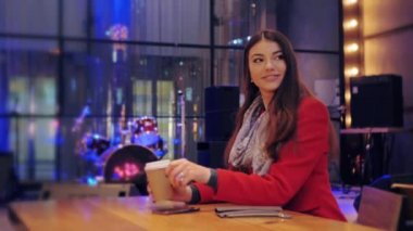 Kırmızı ceketli güzel esmer kadın bir yere bakıyor ve elinde bir fincan kahveyle yemek salonundaki masada otururken gülümsüyor. Kapalı. 