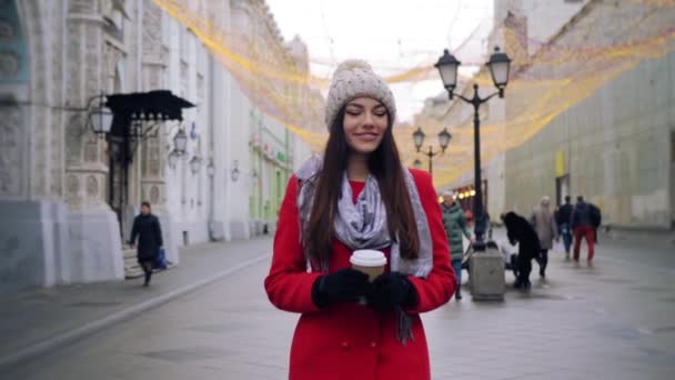 赤いコートの魅力的な女性は屋外で通りを歩いている間にコーヒーを飲んでいます — ストック動画
