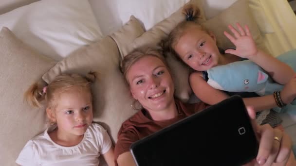 Madre con dos chicas lindas se acuestan en la cama y se comunican usando la tableta — Vídeos de Stock