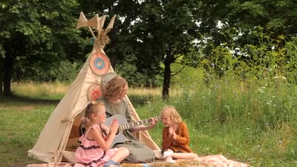 Mam speelt gitaar voor haar twee dochters op het zomerfestival. Wigwam decoratie. — Stockvideo