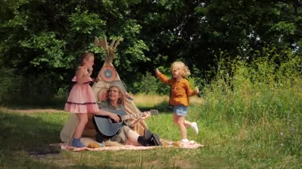 Мати грає на гітарі зі своїми дітьми, зябра танцюють поруч з вігвамом на відкритому повітрі — стокове відео